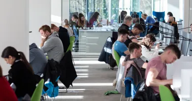 Students sitting at desks at opposite sides of a large bright room. The students are studying and doing work on laptops and personal devices