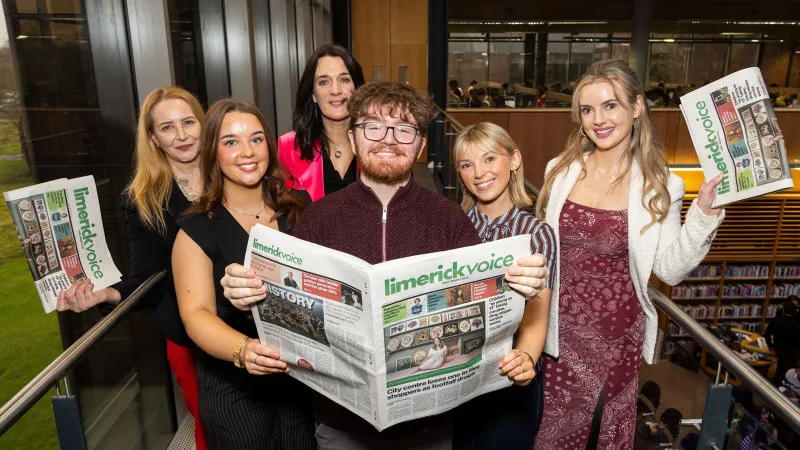 A group of six people in smart dress standing together and holding up newspapers