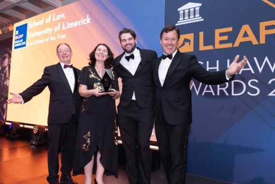 Four people in formal dress standing in front of a yellow and blue screen. One of the people is holding an award.