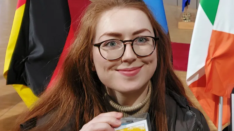 UL graduate Rebecca Zamponi at work in the European Parliament. There are flags behind her and she's holding up a badge with her name on it.