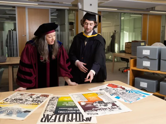 UL graduate Caimin Walsh and UL lecturer Dr Aileen Dillane. Caimin is dressed in a black graduation robe and a black graduation hat. Aileen is dressed in a maroon PhD graduation robe and black hat. They are leaning over a table which has screen printed posters laid out in front of them.