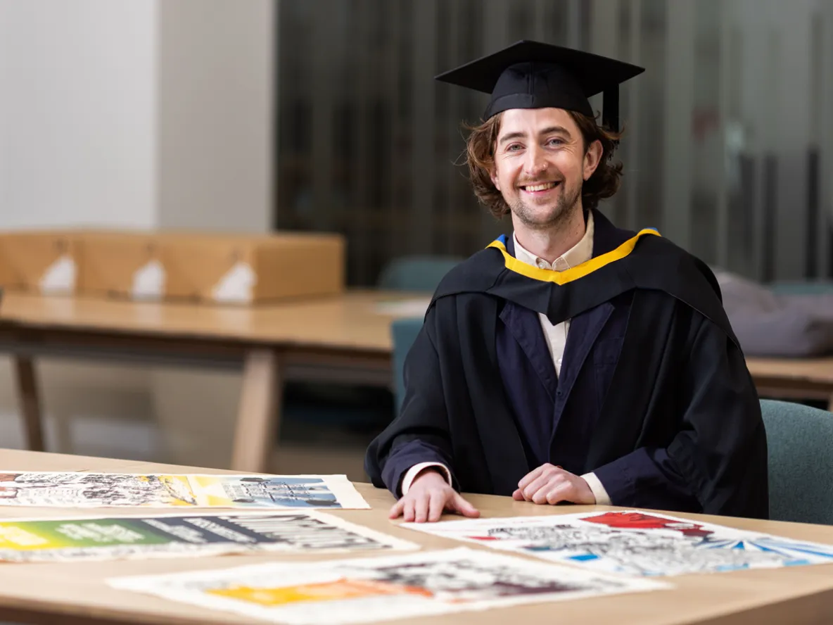 UL graduate Caimin Walsh. He is dressed in a black graduation robe and a black graduation hat. He is seated at a desk and there are screen printed posters on the desk in front of him.