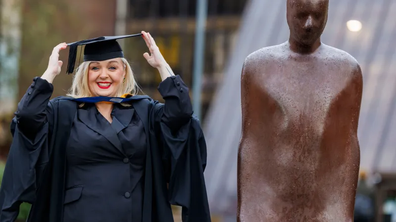 A woman in smart dress and black graduation robes and cap, standing beside a bronze statue of a person. The woman is smiling and holding the cap with both hands.