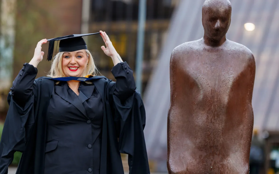 A woman in smart dress and black graduation robes and cap, standing beside a bronze statue of a person. The woman is smiling and holding the cap with both hands.