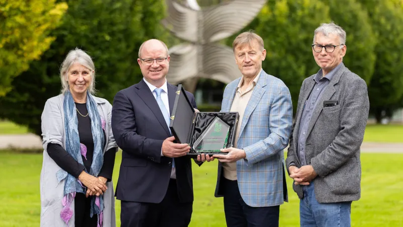 Three men and a woman standing outside in front of a large steel sculpture and green grass. The two men in the middle are holding a glass award in a presentation case. 
