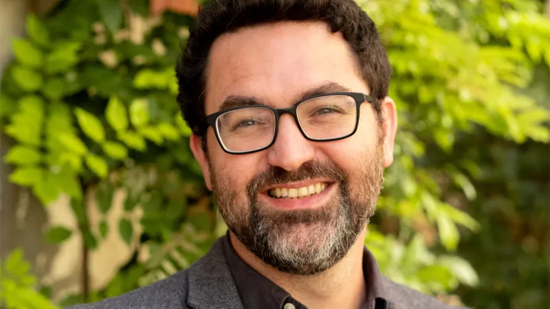 A man with short dark hair and glasses in a grey shirt and jacket standing in front of a green bush