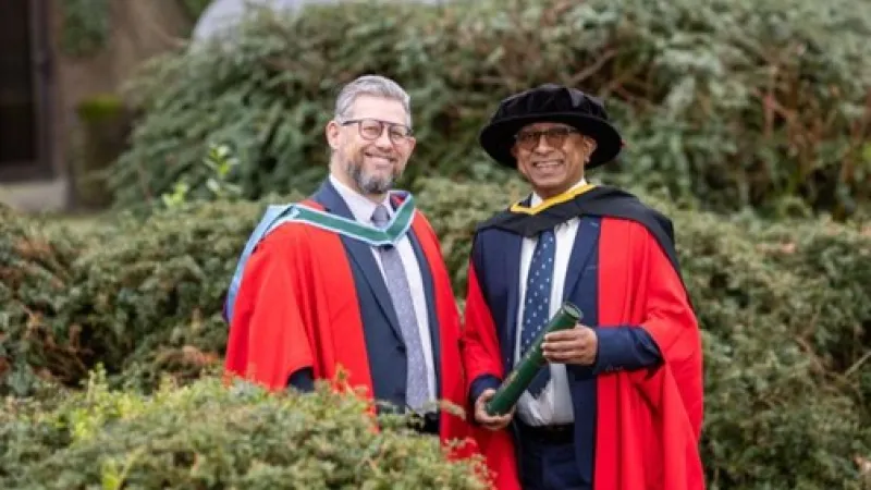 Two men in red academic dress smile at the camera. The man on the right is holding a scroll.
