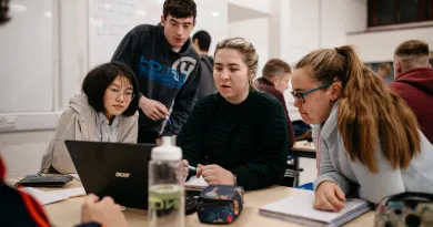 Four young people looking at a laptop on a desk. The group are leaning in towards the laptop with the girl on the right ready to take notes