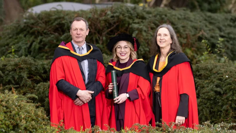 Two women and a man standing among greenery, dressed in red graduation robes. The woman in the middle is also wearing a black graduation cap and holding a green parchment holder